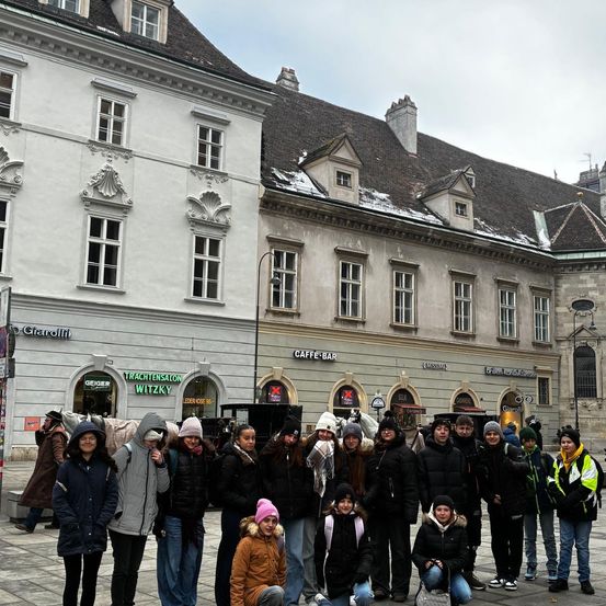 Eine Gruppe von Menschen versammelt sich vor einem Gebäude mit architektonischen Designs und Schnee auf dem Dach. Das Gebäude hat Glasfenster und Schilder für CAFFEE BAR und GEIGER.