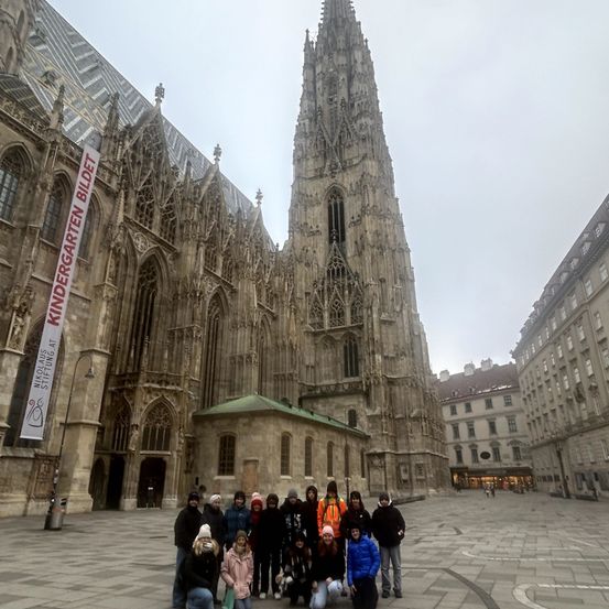 Eine Gruppe von Menschen posiert für ein Foto vor einer großen Kathedrale mit komplizierten architektonischen Details. Ein Banner ist an der Seite des Gebäudes sichtbar.
