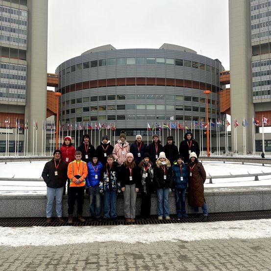 Eine Gruppe von Personen steht vor einem Gebäude mit Fahnen dahinter, einige tragen Mützen und Ausweise, andere sind in Mänteln gekleidet. Der Boden ist schneebedeckt.