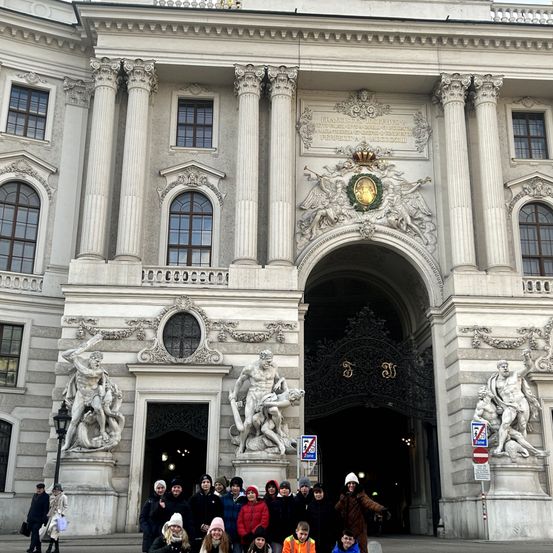 Eine Gruppe von Menschen posiert vor dem Hofburg-Palast in Wien, mit seinen großen Säulen und kunstvollen Statuen. Der Eingang zeichnet sich durch einen Bogen und dekorative Details aus.
