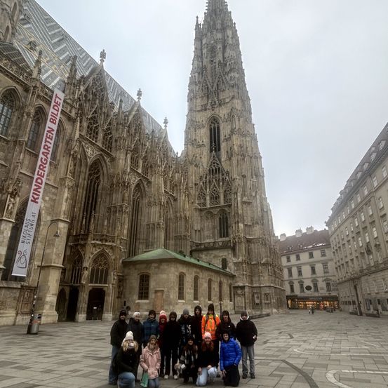 Eine Gruppe von Menschen posiert für ein Foto vor einer gotischen Kathedrale, mit einem Banner, das 'Kindergarten Bildung' steht. Das Wetter ist bewölkt, und es gibt Gebäude an den Seiten.