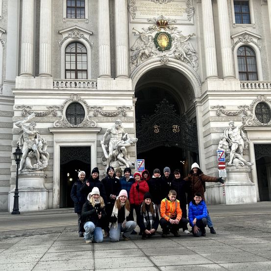 Eine Gruppe von Menschen in Winterkleidung posiert für ein Foto vor der Wiener Staatsoper, einem prächtigen Gebäude mit Statuen und großem Bogeneingang.