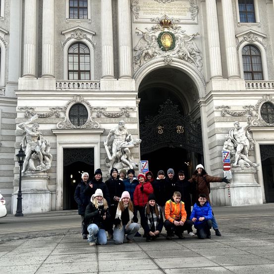 Eine Gruppe von Menschen in Winterkleidung posiert für ein Foto vor dem Hofburg-Palast in Wien. Der Palast zeigt Statuen, einen gewölbten Eingang und dekorative Elemente.