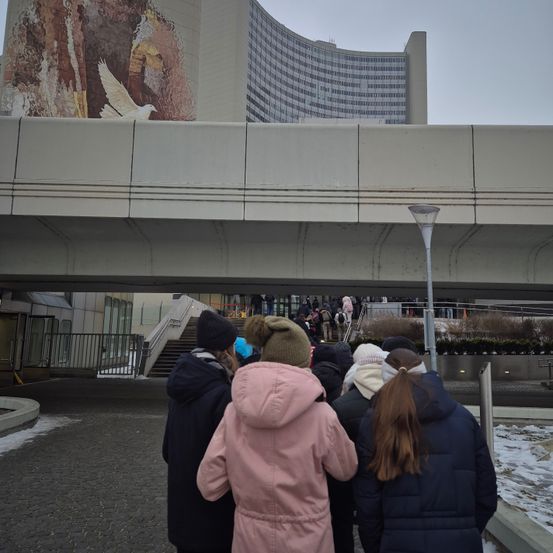 Menschen in Winterkleidung stehen unter einem Überweg und blicken auf ein Gebäude mit einem großen Wandbild einer Taube und eines Baumes. Schnee bedeckt den Boden und ein Lampenpfahl steht in der Nähe.