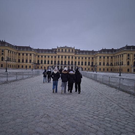 Eine Gruppe von Menschen geht auf einem Kopfsteinpflasterweg vor einem großen, gelben Gebäude mit vielen Fenstern und einem verschneiten Boden.