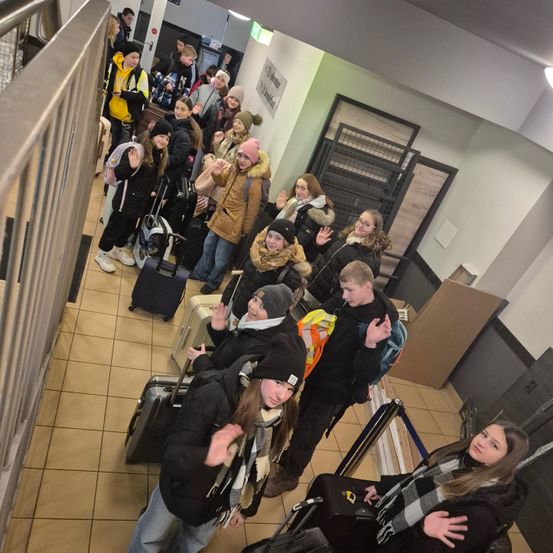 Eine Gruppe von Menschen, wahrscheinlich Schüler, steht in einem Flur und posiert für ein Foto. Sie tragen verschiedene Winterkleidung, Brillen und einige haben Rucksäcke dabei. Gepäck ist umher verstreut, und einige tragen Handschuhe. Ein Metallgeländer und ein Schild sind sichtbar.