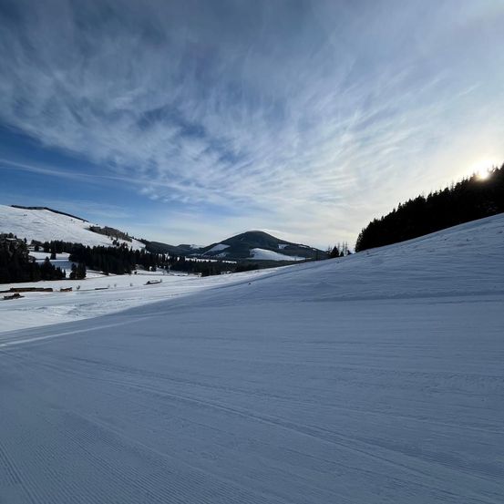 Ein schneebedeckter Berghang mit blauem Himmel und einigen Bäumen in der Ferne. Die Sonne geht am Horizont unter.