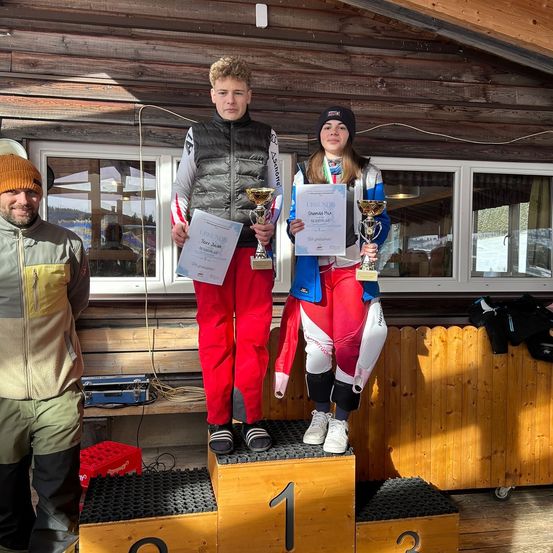 Zwei junge Athleten stehen auf einem Podest und halten Pokale und Urkunden. Der Mann trägt eine schwarze Weste, während die Frau eine blaue Jacke trägt. Hinter ihnen ist eine Holzstruktur und ein Fenster zu sehen.