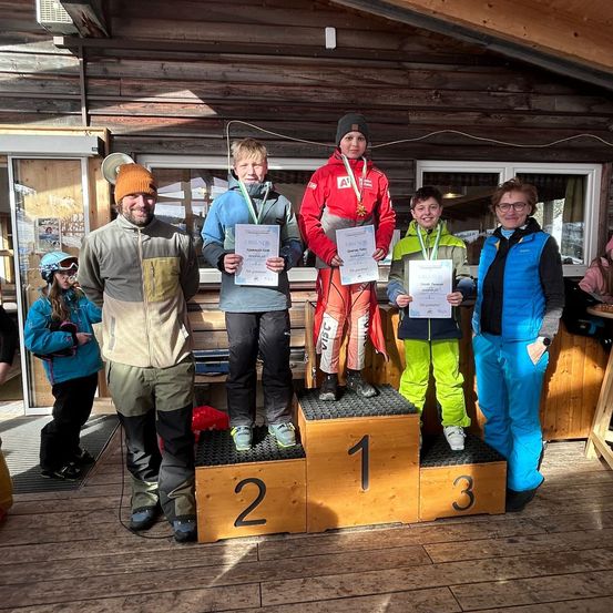 Fünf Personen stehen auf einem Podest mit Medaillen und Urkunden, wahrscheinlich zur Feier eines Skiwettbewerbs. Drei links tragen Skiausrüstung, während zwei rechts Jacken und Brillen tragen. Dahinter steht eine Holzstruktur.