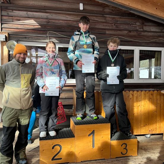 Drei Kinder stehen auf einem Podium mit Medaillen um den Hals. Sie lächeln und halten Urkunden in den Händen. Sie stehen auf einem hölzernen Podest mit den Nummern 1, 2 und 3. Hinter ihnen ist ein Holzzaun.