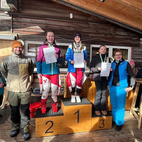 Fünf Personen stehen auf einem Podium mit drei Stufen. Sie lächeln und halten Urkunden in den Händen. Auf dem Podium stehen die Zahlen 1, 2 und 3. Die Person auf der ersten Stufe trägt eine Medaille.