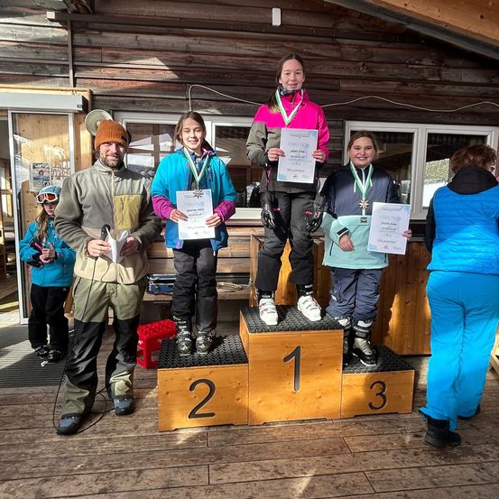 Eine Gruppe von Menschen, darunter zwei junge Mädchen, steht auf einem Podium. Sie halten Urkunden in den Händen und scheinen zu feiern. Hinter ihnen steht ein Mann mit einem Mikrofon und einer weiteren Urkunde. Sie befinden sich in einer Holzhütte.