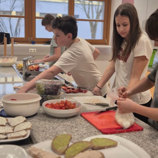Vier Kinder bereiten Essen in einer Küche zu. Eines schneidet Tomaten, ein anderes handhabt eine Plastiktüte, und zwei andere konzentrieren sich auf Aufgaben in der Nähe einer Spüle mit einem Wasserhahn. Schüsseln, Teller und ein Schneidebrett stehen auf der Arbeitsplatte.