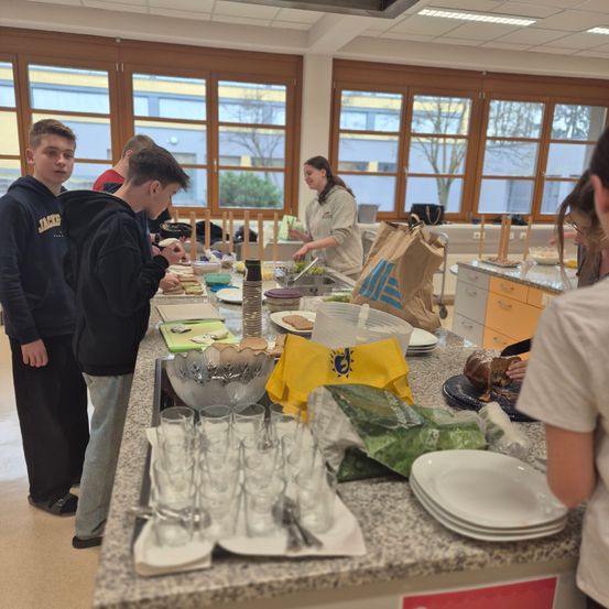 Eine Gruppe junger Leute bereitet in einer Küche Essen zu. Eine Person schneidet Gemüse, während andere Essen auf Teller servieren. Auf dem Tresen befinden sich Gläser, Teller und Schüsseln.
