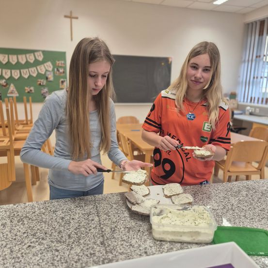 Zwei Mädchen bereiten in einem Klassenzimmer Sandwiches zu. Eine benutzt ein Messer, während die andere ein Sandwich hält. Ein Tafel mit Dekorationen ist im Hintergrund.