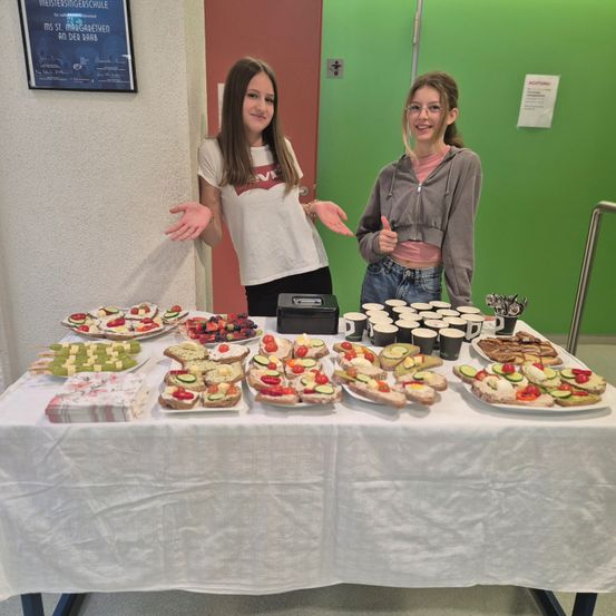 Zwei Frauen stehen hinter einem Tisch voller Sandwiches, Früchten und Getränken. Eine Frau trägt ein weißes T-Shirt und die andere eine graue Hoodie. Sie lächeln und posieren für ein Foto.