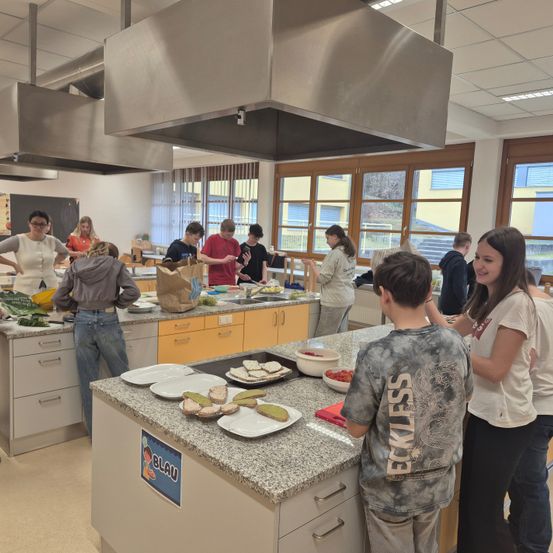 Schüler in einer Klassenküche bereiten gemeinsam Essen zu. Sie stehen um einen Tisch mit Tellern voller Essen, ein Schüler hält ein Messer. Die Küche hat eine Arbeitsplatte, Schränke und Fenster mit gelben Rahmen.