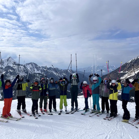 Eine Gruppe von Menschen in Winterkleidung posiert für ein Foto auf einem verschneiten Berggipfel. Sie halten Skistöcke und stehen auf Skiern. Der Hintergrund zeigt schneebedeckte Berge.