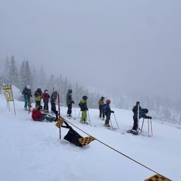 Eine Gruppe von Skifahrern in verschiedenen Skiausrüstungen wartet in einer Reihe auf einem schneebedeckten Hang mit Tannen im Hintergrund.