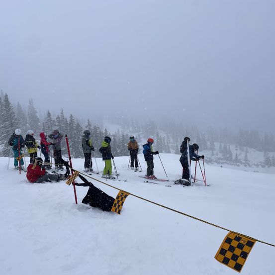 Skifahrer und Snowboarder warten in einer Schlange auf einem verschneiten Berg. Eine Person ist gefallen und andere beobachten.