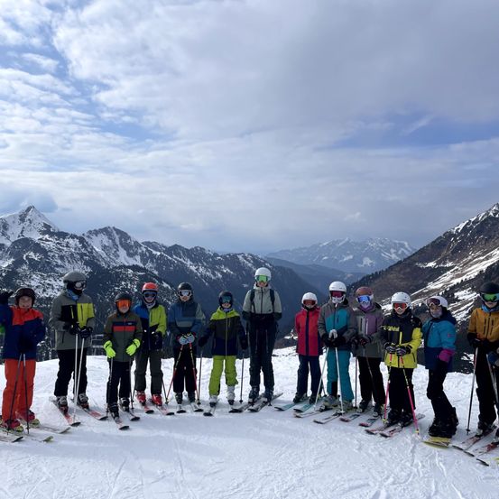 Eine Gruppe von Skifahrern steht auf einem verschneiten Berg und trägt Winterausrüstung. Sie alle lächeln und posieren für ein Foto. Hinter ihnen sind schneebedeckte Berge unter einem teilweise bewölkten Himmel sichtbar.