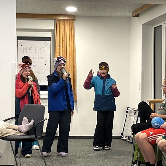 Vier Kinder in Winterkleidung stehen in einem Raum und singen in Mikrofone. Ein Whiteboard ist hinter ihnen sichtbar.