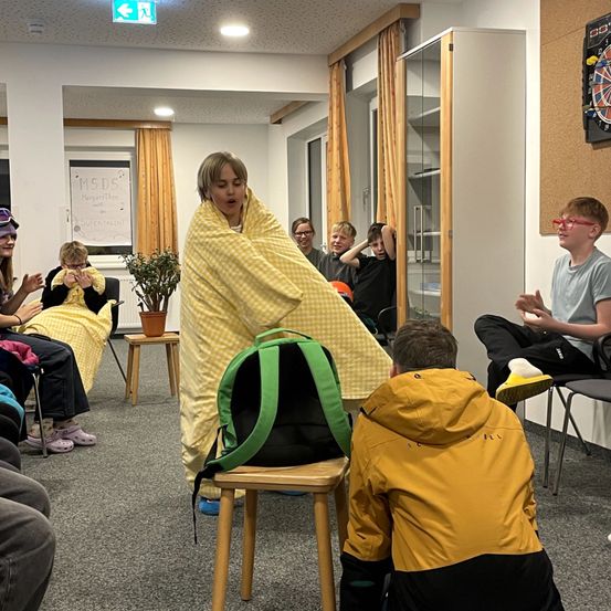 Ein Klassenzimmer mit sitzenden Schülern, eine in Gelb, die sich eine Decke umwickelt, ein anderer kniet vor ihr, ein Rucksack auf einem Hocker in der Nähe.