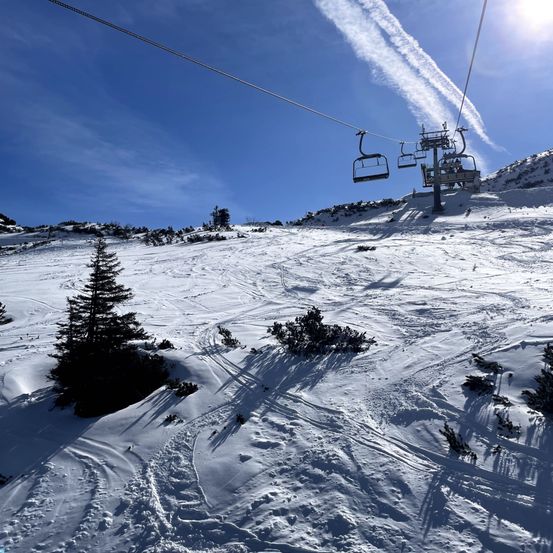 Eine Skiliftkabine schwebt über einem verschneiten Berghang mit Skispuren und immergrünen Bäumen. Der Himmel ist blau mit einigen Wolken und einer strahlenden Sonne.