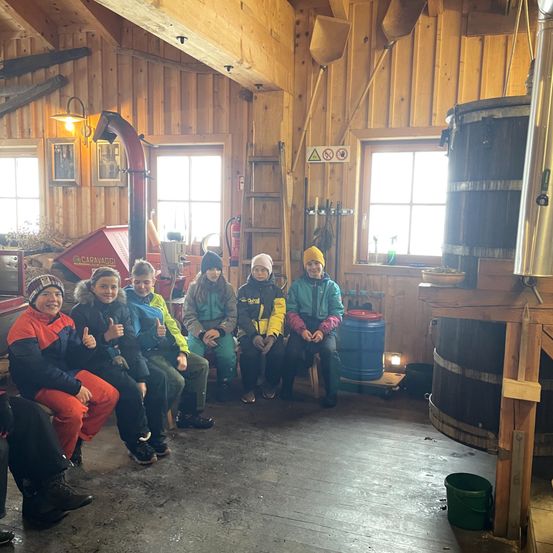 Eine Gruppe von Kindern in Winterkleidung sitzt auf einer Bank in einem Raum. Ein Feuerlöscher ist an der Wand angebracht, und eine Holzleiter steht in der Nähe des Fensters. Ein großer hölzerner Fass steht in der Nähe der Wand, und eine Lampe hängt an der Decke.