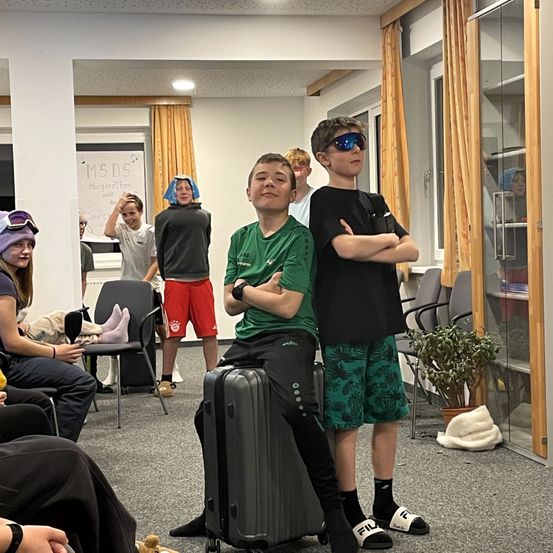 Mehrere junge Menschen stehen in einem Raum. Ein Junge sitzt auf einem Koffer. Ein anderer Junge lehnt sich mit verschränkten Armen an eine Wand. Hinter ihnen befinden sich Stühle und eine Tafel.