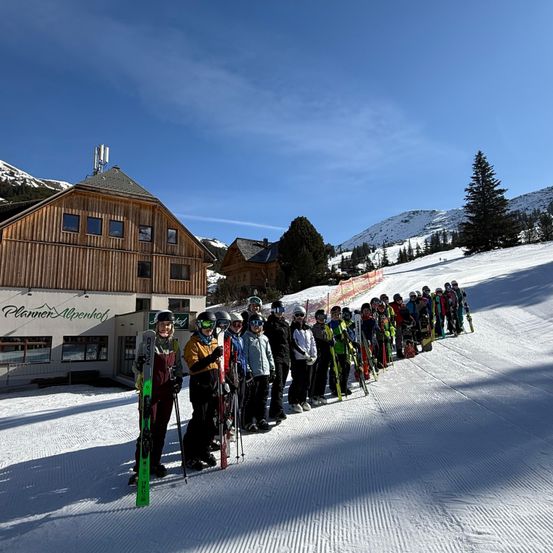 Eine Gruppe von Skifahrern steht auf einem verschneiten Hang, mit einem Gebäude und Bergen im Hintergrund.