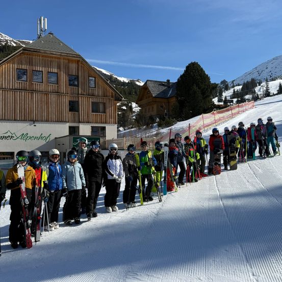 Eine Gruppe von Menschen in Winterausrüstung steht in einer Reihe auf einem verschneiten Hang, mit einem Gebäude im Hintergrund.