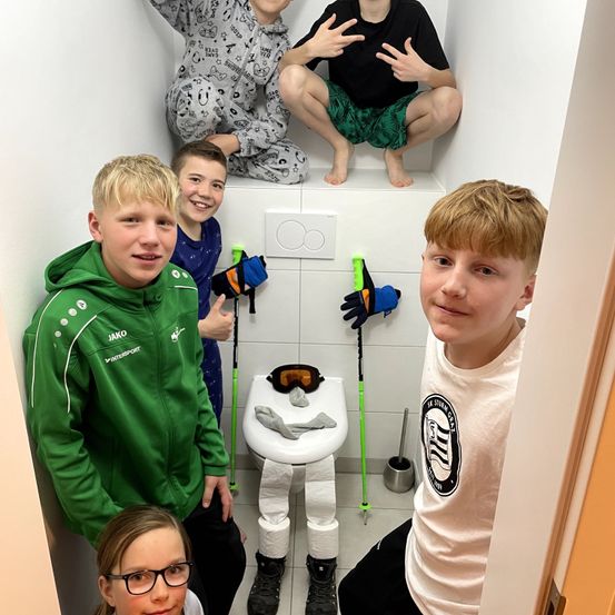 Eine Gruppe junger Jungen und ein Mädchen posiert in einem Badezimmer. Einige von ihnen sitzen auf einem Toilettensitz mit Toilettenpapier und Handschuhen. Sie alle scheinen für ein Foto zu lächeln.