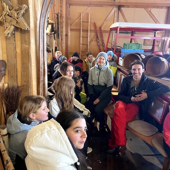 Eine Gruppe von Menschen, einschließlich Kindern, versammelt sich drinnen. Sie tragen Winterkleidung und scheinen auf einer Tour zu sein. Ein Mann sitzt auf einer Bank und lächelt.