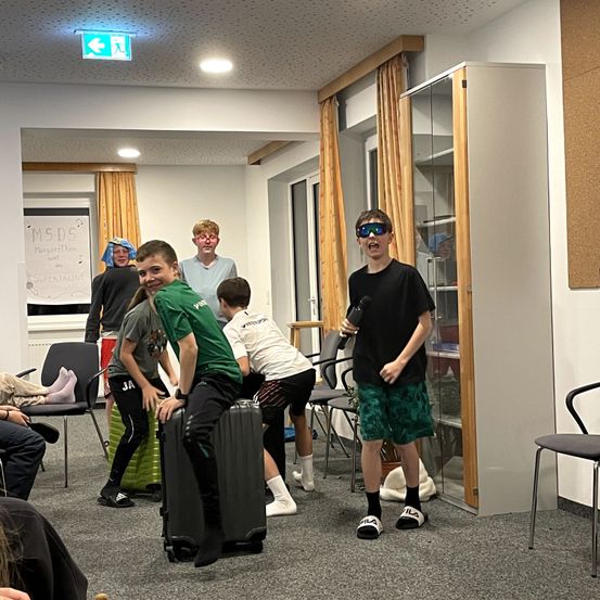 Mehrere Kinder in einem Raum, eines trägt eine Sonnenbrille und hält ein Mikrofon. Ein Koffer liegt auf dem Boden und ein Schild mit der Aufschrift MSDS und Margarethen ist an der Wand angebracht.