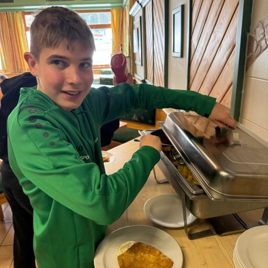 Ein Junge in einem grünen Pullover serviert sich selbst Essen von einem Buffet. Er lächelt und hält einen Löffel.