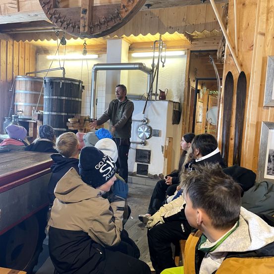 Ein Mann steht in einem Raum mit Holzwänden und erklärt etwas einer Gruppe von Kindern, die vor ihm sitzen. Im Hintergrund befinden sich große Holzfässer und verschiedene Werkzeuge.