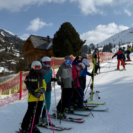 Eine Gruppe von Kindern in Skiausrüstung steht auf einem verschneiten Hang. Sie bereiten sich auf eine Skistunde vor. Ein Holzhaus ist im Hintergrund zu sehen.