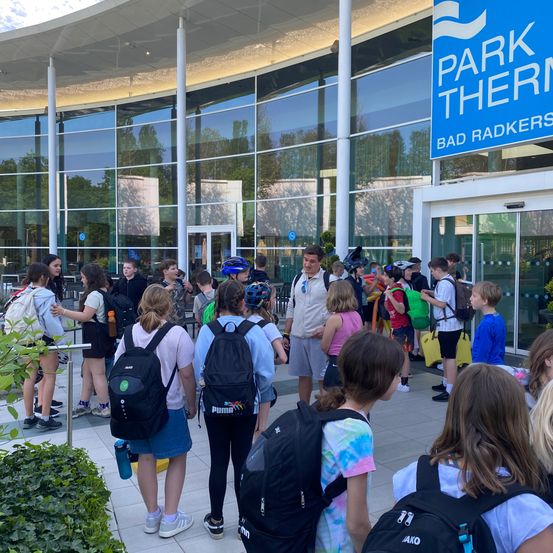 Eine Gruppe von Schülern und Lehrern steht vor einem Gebäude mit Glaswänden, beschriftet mit Park Therm Bad Radkers. Einige tragen Rucksäcke, und einige halten Helme. Bäume und Büsche sind in der Nähe.