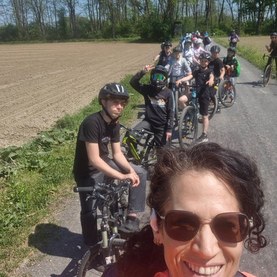 Eine Gruppe von Radfahrern mit Helmen fährt auf einer Feldweg, ein Feld ist neben ihnen, eine Frau macht ein Selfie mit ihnen.