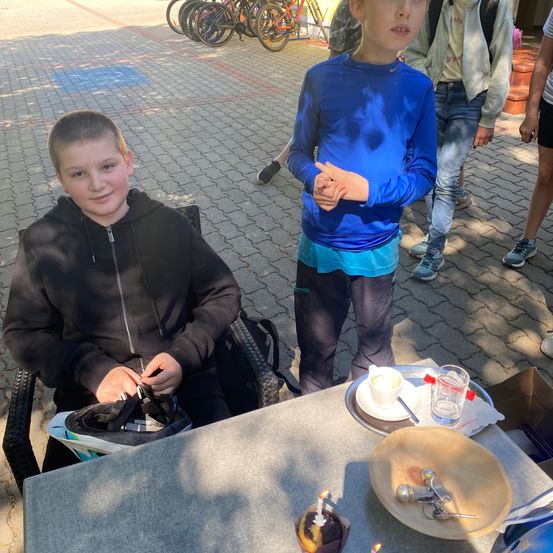 Zwei Jungen sind draußen. Einer sitzt auf einem Stuhl und zieht seinen Helm ab, während der andere steht und lächelt. Sie sind in der Nähe eines Tisches mit einer Tasse und einem Teller. Im Hintergrund sind Fahrräder geparkt.