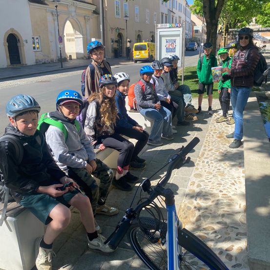 Eine Gruppe von Kindern mit Helmen sitzt auf einer Bank, ein Kind hält ein Handy, und ein Erwachsener steht neben ihnen mit einem Buch.