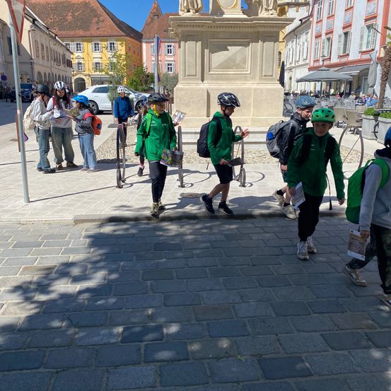 Eine Gruppe von Kindern und Erwachsenen in grünen Jacken und Helmen geht auf einer Kopfsteinpflasterstraße. Hinter ihnen steht ein Denkmal. Einige von ihnen tragen Bücher. Im Hintergrund sind Gebäude und geparkte Autos.