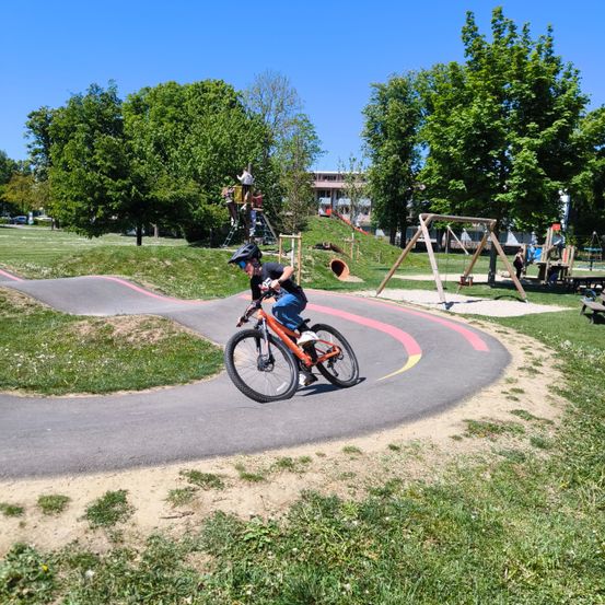 Ein Radfahrer fährt ein orangefarbenes Fahrrad auf einem gewundenen Weg in einem Park mit Bäumen und Spielgeräten.