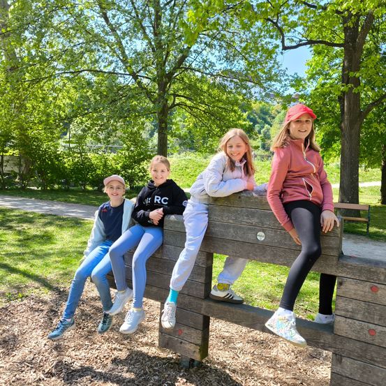Vier Mädchen sitzen auf einer Holzbank in einem Park, lächeln und posieren für ein Foto. Die Bank steht auf dem Boden in einem Grasbereich. Das Gebiet ist von Bäumen und einer Bank auf der rechten Seite umgeben. Das Mädchen auf der rechten Seite trägt eine Mütze.