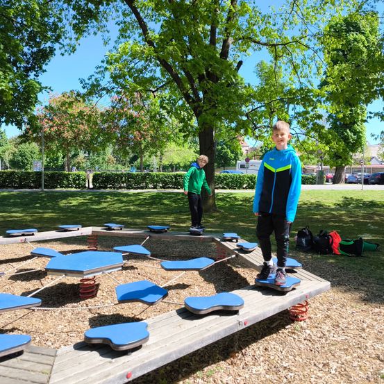 Zwei Jungen spielen auf einer kreisförmigen blauen Federplattform in einem Park, einer trägt einen blauen und der andere einen grünen Pullover.