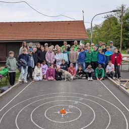 Eine Gruppe von Menschen posiert vor einem Gebäude mit einem roten Dach. Sie stehen um einen Kreis mit Zahlen und einer Zielscheibe. Die Umgebung ist draußen.