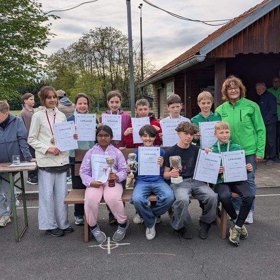 Mehrere Kinder und eine Frau halten Zertifikate bei einer Veranstaltung im Freien. Sie sitzen auf einer Bank. Sie tragen Turnschuhe. Dahinter befindet sich ein Gebäude mit Dach und offener Tür.