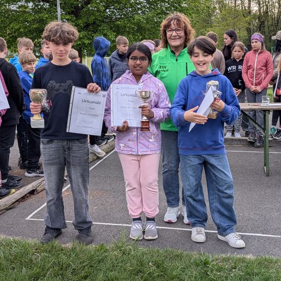 Drei junge Jungen und ein Mädchen, alle mit Trophäen und Zertifikaten in der Hand, stehen in einem Außenbereich. Dahinter stehen mehrere Menschen und beobachten.