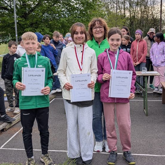 Drei Kinder stehen vor einem Tisch und halten Urkunden für den zweiten Platz bei einer Veranstaltung. Sie sind von Zuschauern in einem Park umgeben.